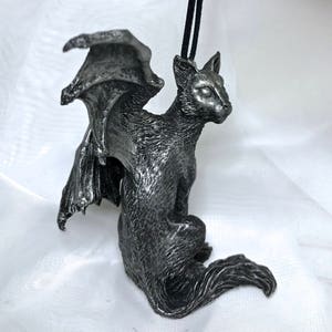 May include: A silver-colored figurine of a cat with bat wings. The cat is sitting with its tail curled around its legs. The figurine is made of a textured material and has a realistic appearance.