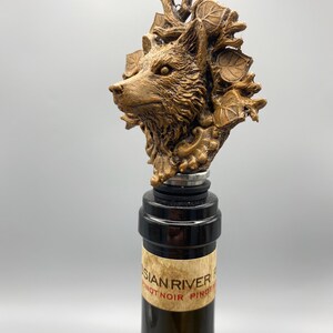 Op de afbeelding: Een gouden wolfskop flessenstop met bladeren en takken. De stop zit op een wijnfles met een etiket waarop "Russian River Pinot Noir" staat.
