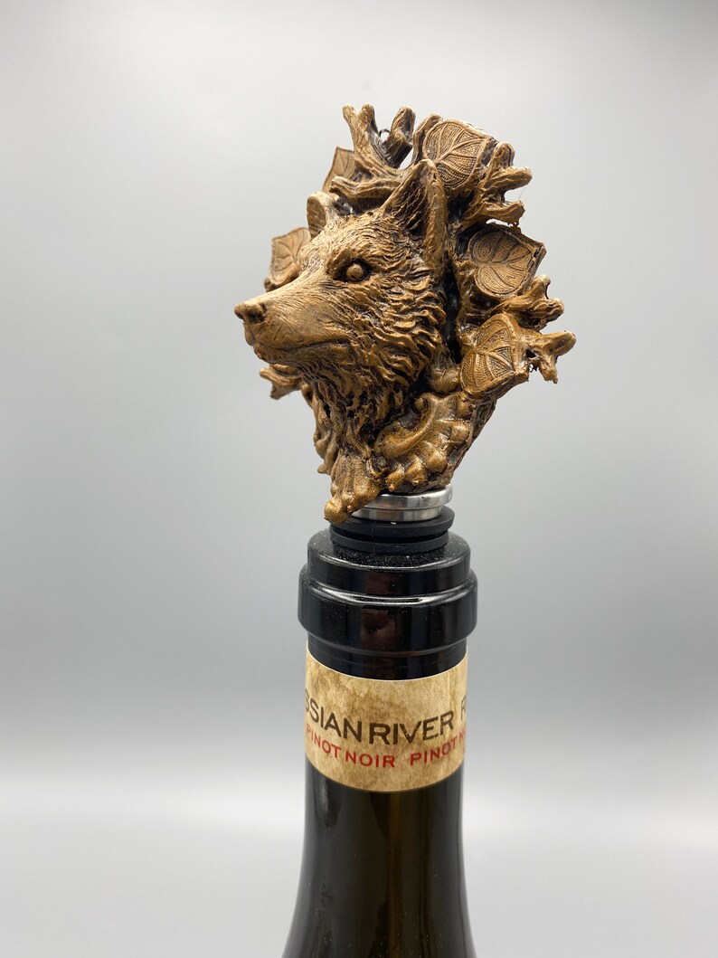 Op de afbeelding: Een gouden wolfskop flessenstop met bladeren en takken. De stop zit op een wijnfles met een etiket waarop "Russian River Pinot Noir" staat.