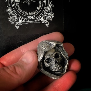 May include: A silver-colored skull wearing a hooded cloak. The skull has detailed features and is set against a black background.