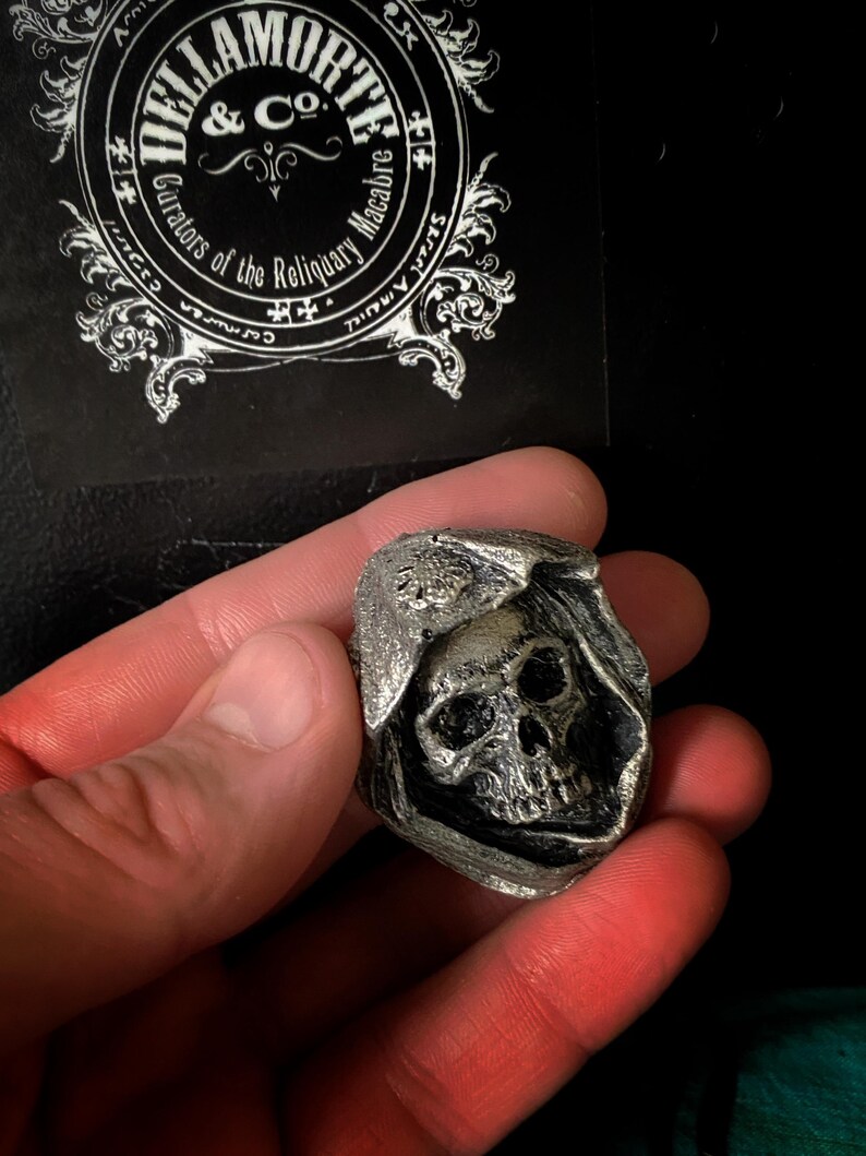 May include: A silver-colored skull wearing a hooded cloak. The skull has detailed features and is set against a black background.