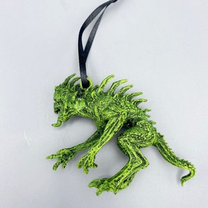 May include: Green monster ornament with a black string for hanging. The monster has a spiky back, sharp teeth, and a long tail.
