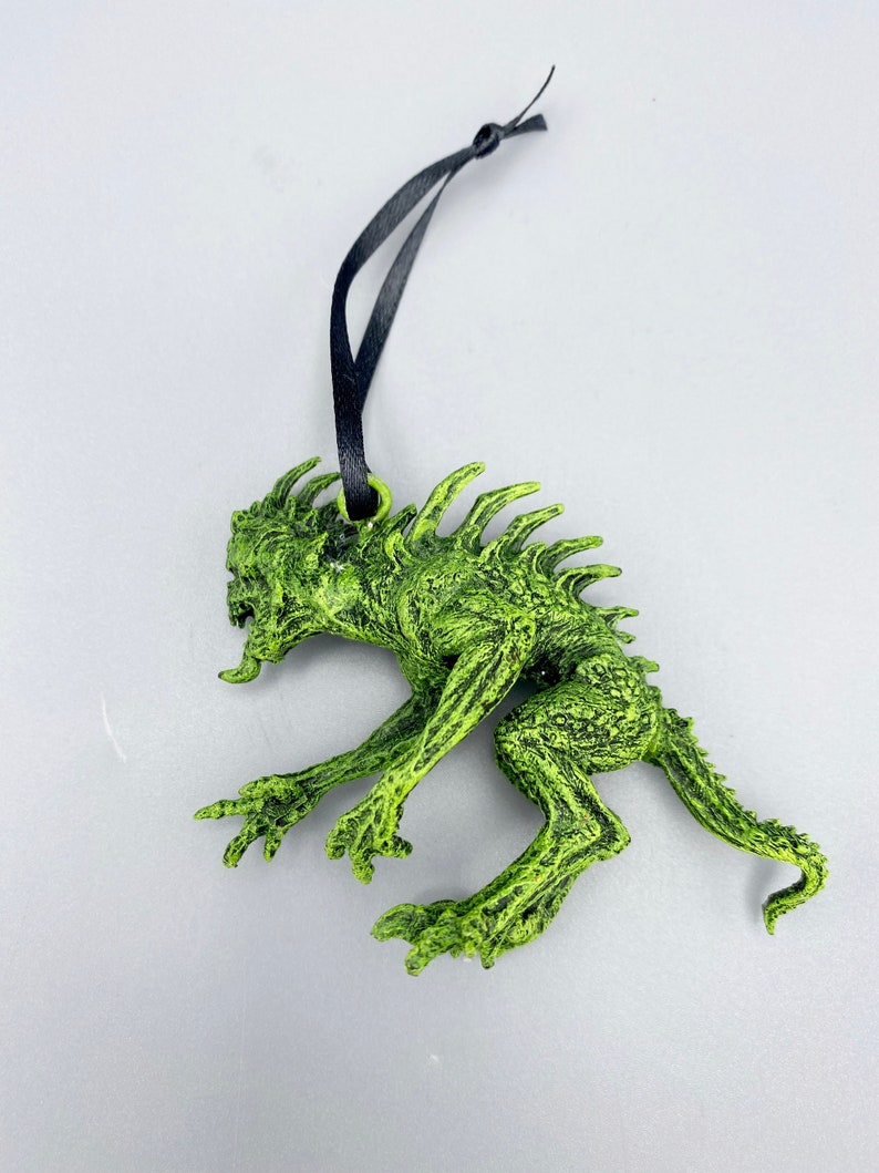May include: Green monster ornament with a black string for hanging. The monster has a spiky back, sharp teeth, and a long tail.