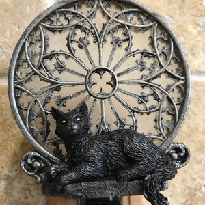 Peut inclure: Une veilleuse en forme de chat noir avec un cadre en m&eacute;tal argent&eacute;. Le cadre est con&ccedil;u pour ressembler &agrave; une fen&ecirc;tre en vitrail. Le chat est assis sur un rebord avec sa queue enroul&eacute;e autour de son corps.