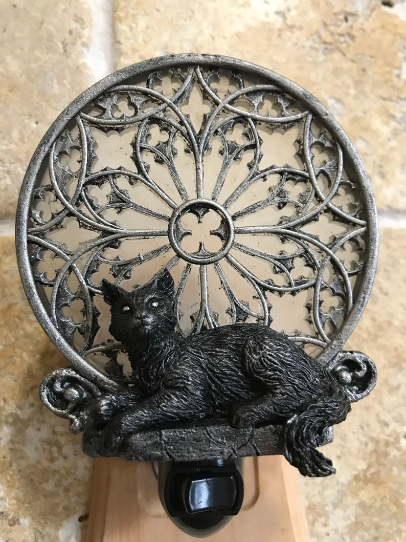 Peut inclure: Une veilleuse en forme de chat noir avec un cadre en m&eacute;tal argent&eacute;. Le cadre est con&ccedil;u pour ressembler &agrave; une fen&ecirc;tre en vitrail. Le chat est assis sur un rebord avec sa queue enroul&eacute;e autour de son corps.