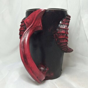 May include: A black and red ceramic mug with a dragon-shaped handle. The mug has a textured surface and a stainless steel rim.