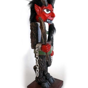 May include: A red and black wooden figurine of a devil with horns, a beard, and a stick in its hand. The figurine is wearing chains and is standing on a wooden base.