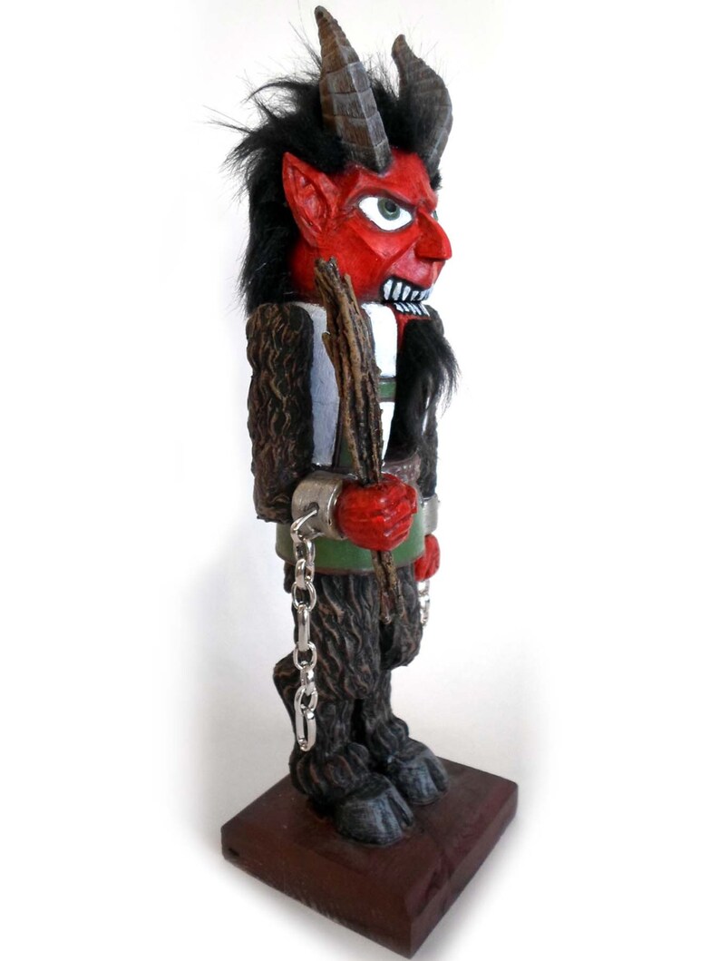 May include: A red and black wooden figurine of a devil with horns, a beard, and a stick in its hand. The figurine is wearing chains and is standing on a wooden base.