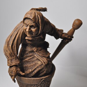 May include: A brown ceramic figurine of a woman with a long, flowing robe and a pointed hat. She is holding a long, thin stick in her right hand. The figurine is standing on a base that is shaped like a basket.