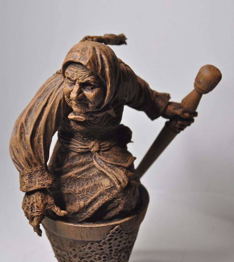 May include: A brown ceramic figurine of a woman with a long, flowing robe and a pointed hat. She is holding a long, thin stick in her right hand. The figurine is standing on a base that is shaped like a basket.