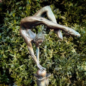 May include: A bronze sculpture of a woman performing a handstand on a ball. The sculpture is set against a backdrop of green foliage.