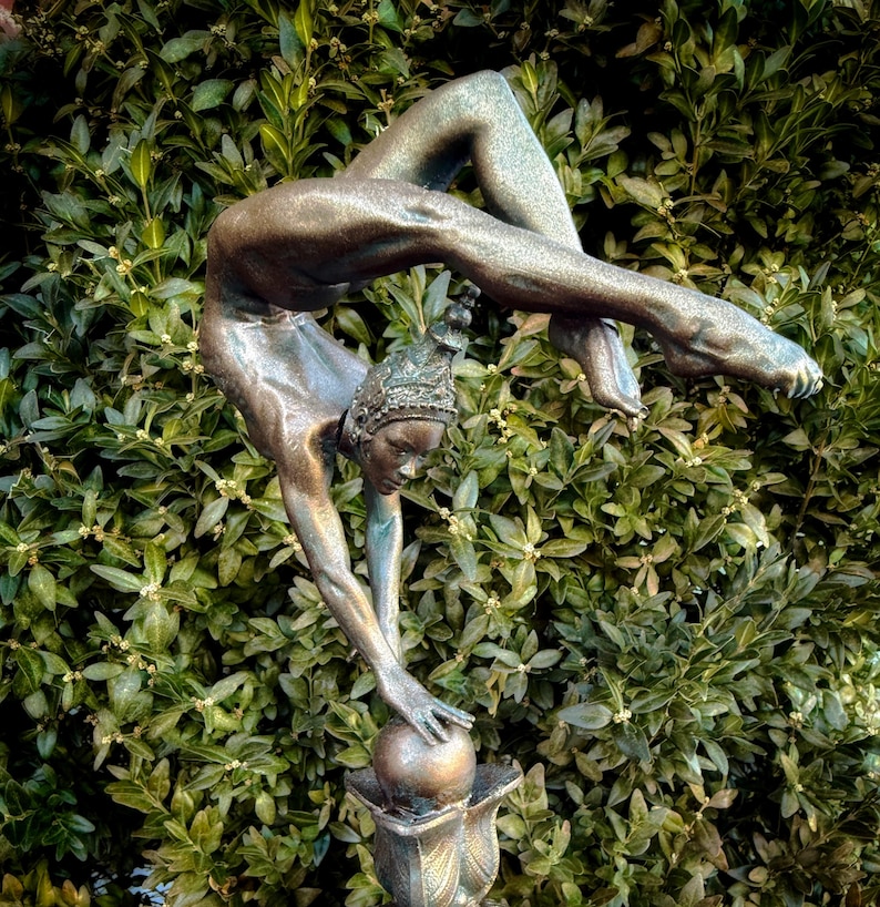 May include: A bronze sculpture of a woman performing a handstand on a ball. The sculpture is set against a backdrop of green foliage.