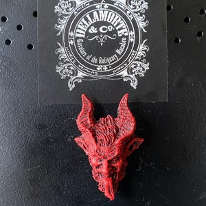 May include: Red resin sculpture of a devil's head with horns and a beard. The sculpture is a small, detailed piece with a textured surface.