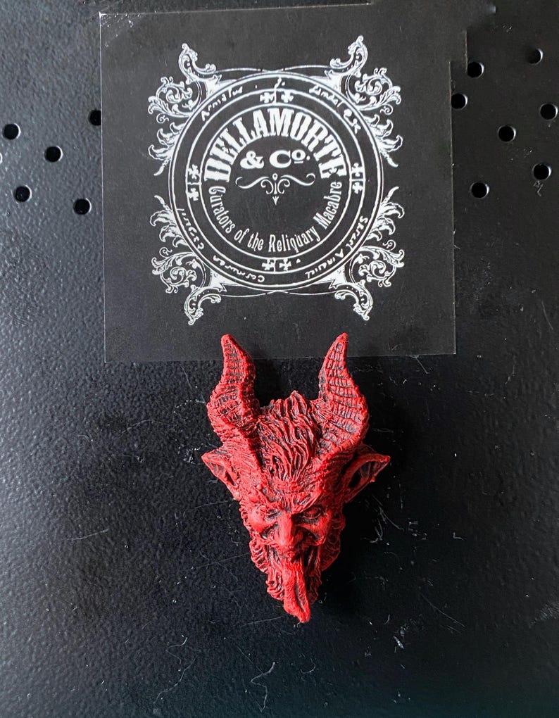 May include: Red resin sculpture of a devil's head with horns and a beard. The sculpture is a small, detailed piece with a textured surface.