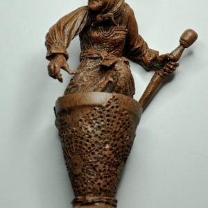 May include: A brown ceramic figurine of a woman holding a wooden pestle. The woman is wearing a long brown dress and a brown head covering. The figurine is sitting in a brown ceramic mortar.