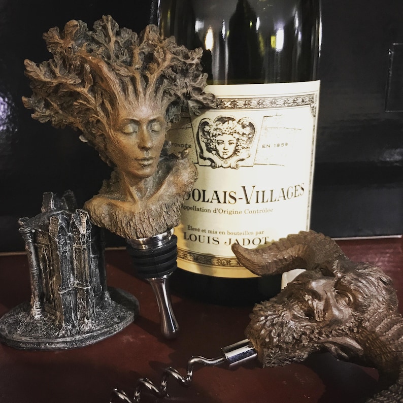 May include: Three decorative bottle stoppers, one featuring a woman's face with tree branches for hair, another with a gothic-style castle, and the third with a horned, bearded creature. The stoppers are made of a brown, textured material. The bottle stoppers are displayed on a dark surface.