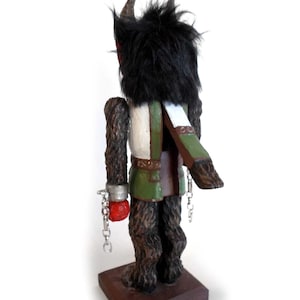 May include: A wooden figurine of a creature with black fur, horns, and a green and white outfit. The creature is chained at the wrists and has a red hand. The figurine is standing on a wooden base.