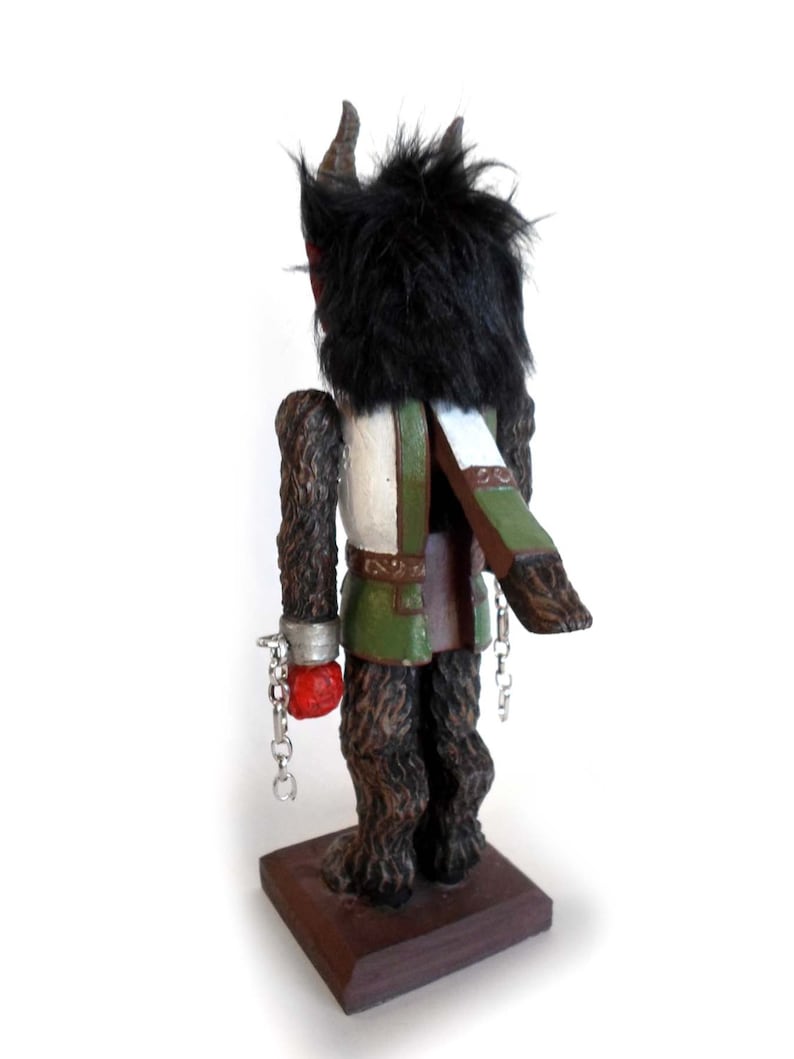 May include: A wooden figurine of a creature with black fur, horns, and a green and white outfit. The creature is chained at the wrists and has a red hand. The figurine is standing on a wooden base.