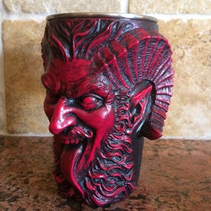May include: A red and black ceramic mug shaped like a demon's head with a long tongue sticking out. The mug has a detailed design with a beard, horns, and a furrowed brow.