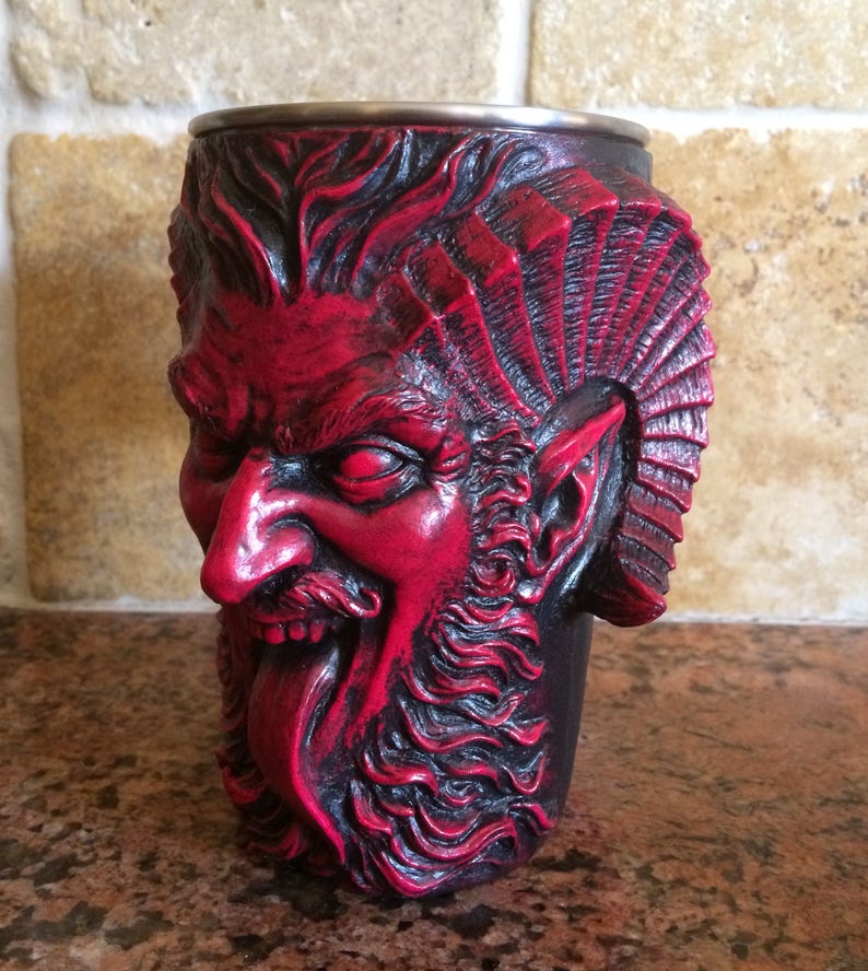May include: A red and black ceramic mug shaped like a demon's head with a long tongue sticking out. The mug has a detailed design with a beard, horns, and a furrowed brow.