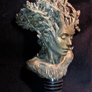 May include: A bronze bottle stopper featuring a detailed sculpture of a woman's face with her hair flowing out in a swirling pattern. The stopper is designed to fit a standard wine bottle.