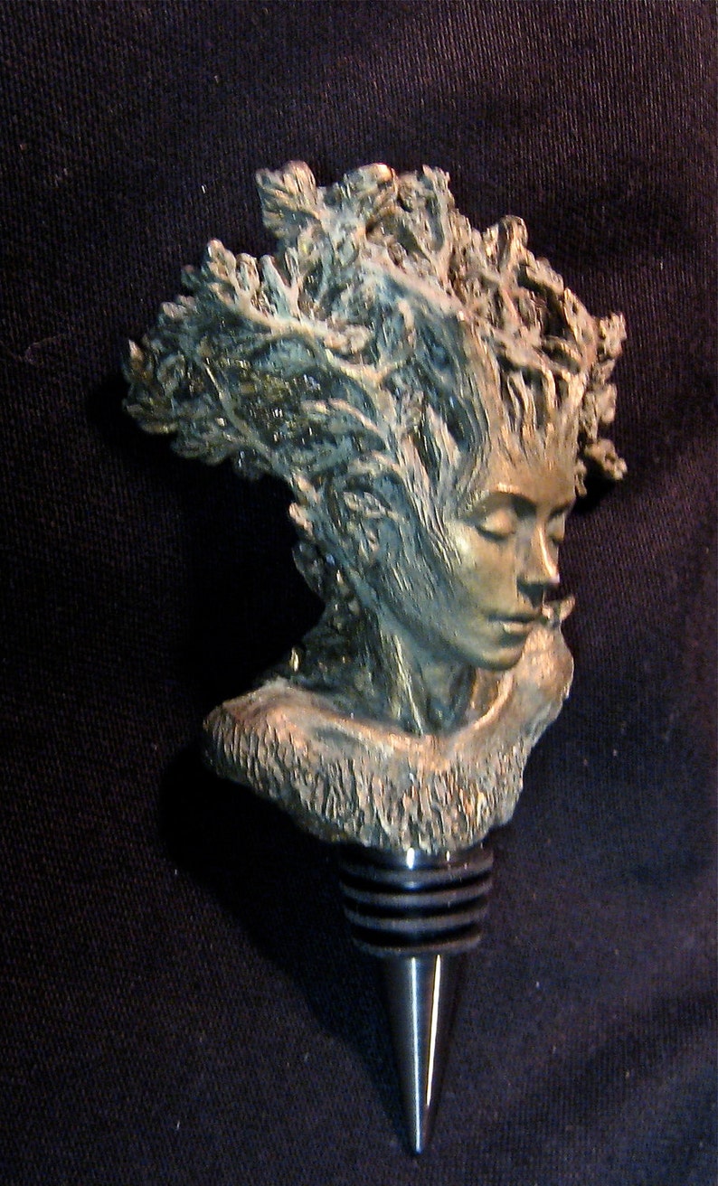 May include: A bronze bottle stopper featuring a detailed sculpture of a woman's face with her hair flowing out in a swirling pattern. The stopper is designed to fit a standard wine bottle.