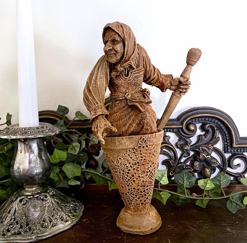 May include: A wooden figurine of a woman holding a wooden spoon, standing in a wooden mortar and pestle. The figurine is painted in shades of brown and has a detailed face.