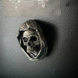 May include: A silver skull with a hooded cloak, the skull has a detailed design and is a decorative item.