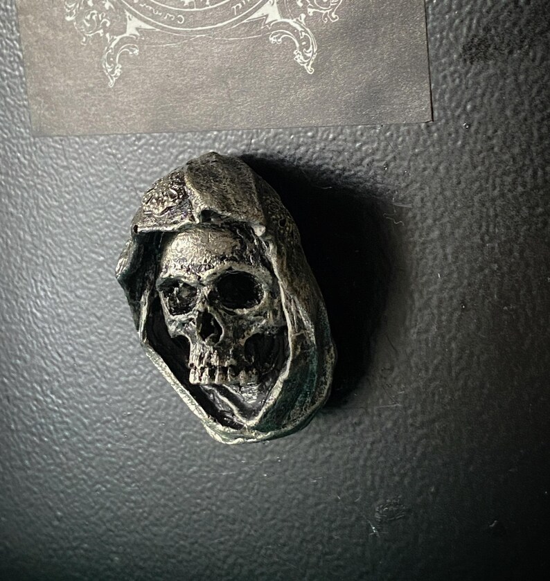 May include: A silver skull with a hooded cloak, the skull has a detailed design and is a decorative item.