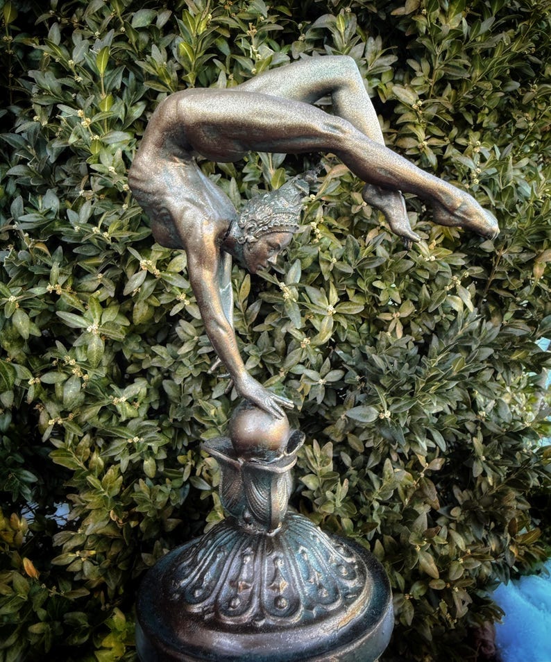May include: A bronze sculpture of a woman performing a handstand on a ball. The sculpture is on a decorative base with intricate details.