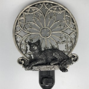 Peut inclure: Une veilleuse argent&eacute;e avec une figurine de chat noir assise sur un rebord de pierre. La veilleuse pr&eacute;sente un motif d&eacute;coratif circulaire en vitrail.
