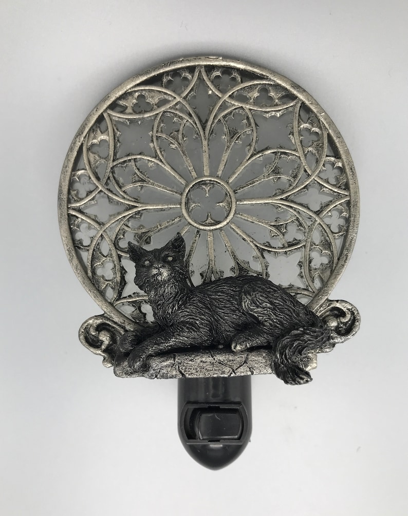 Peut inclure: Une veilleuse argent&eacute;e avec une figurine de chat noir assise sur un rebord de pierre. La veilleuse pr&eacute;sente un motif d&eacute;coratif circulaire en vitrail.