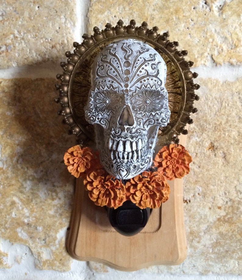 May include: A white sugar skull with gold and black details, surrounded by a gold halo and orange marigold flowers. The skull is attached to a wooden light switch plate.