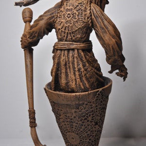 May include: A brown ceramic figurine of a woman wearing a hooded cloak and holding a staff. The figurine is standing on a brown ceramic base with a decorative pattern.