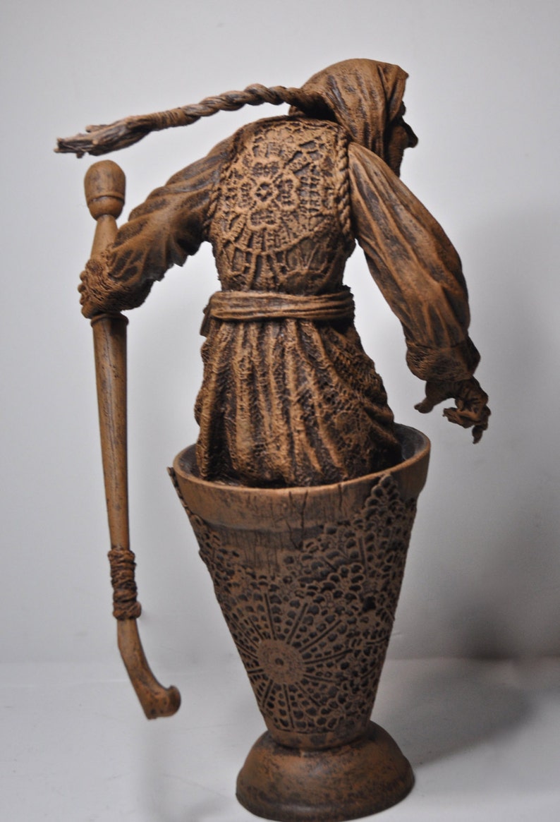 May include: A brown ceramic figurine of a woman wearing a hooded cloak and holding a staff. The figurine is standing on a brown ceramic base with a decorative pattern.