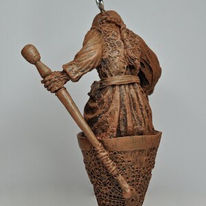 May include: A brown ceramic ornament of a person holding a staff, standing in a cup-shaped base with a decorative pattern. The ornament is hanging from a black cord.