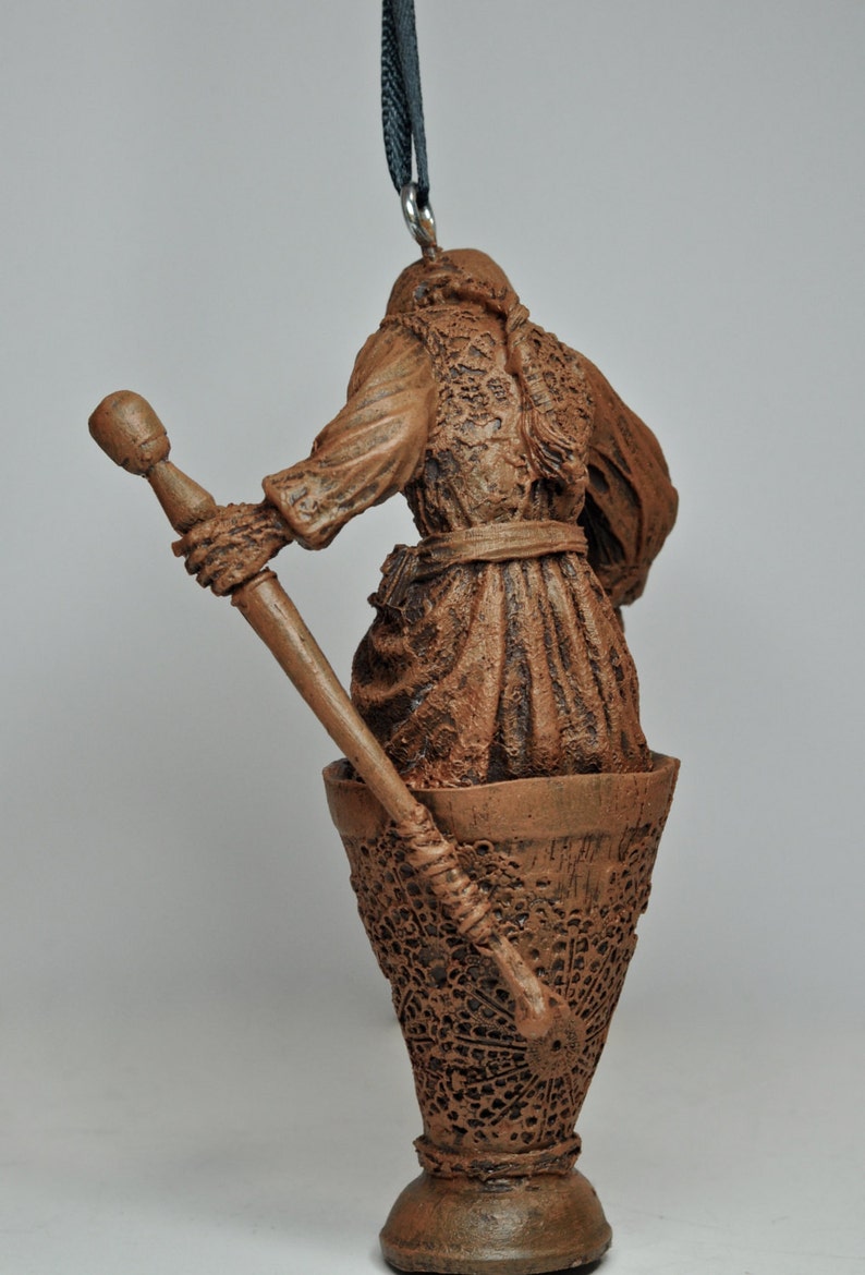 May include: A brown ceramic ornament of a person holding a staff, standing in a cup-shaped base with a decorative pattern. The ornament is hanging from a black cord.