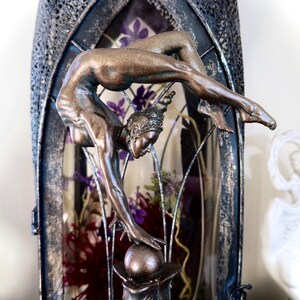 May include: A bronze-colored sculpture of a woman in a contortionist pose, balancing on a sphere. The sculpture is displayed inside a decorative glass cage with dried flowers.