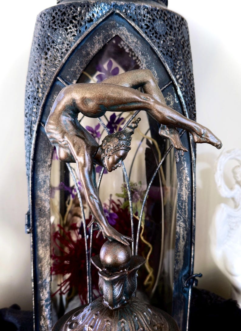 May include: A bronze-colored sculpture of a woman in a contortionist pose, balancing on a sphere. The sculpture is displayed inside a decorative glass cage with dried flowers.