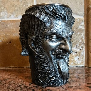 May include: A silver metal drinking cup shaped like a demon's head with a long tongue sticking out. The cup has a detailed design with a beard and horns.