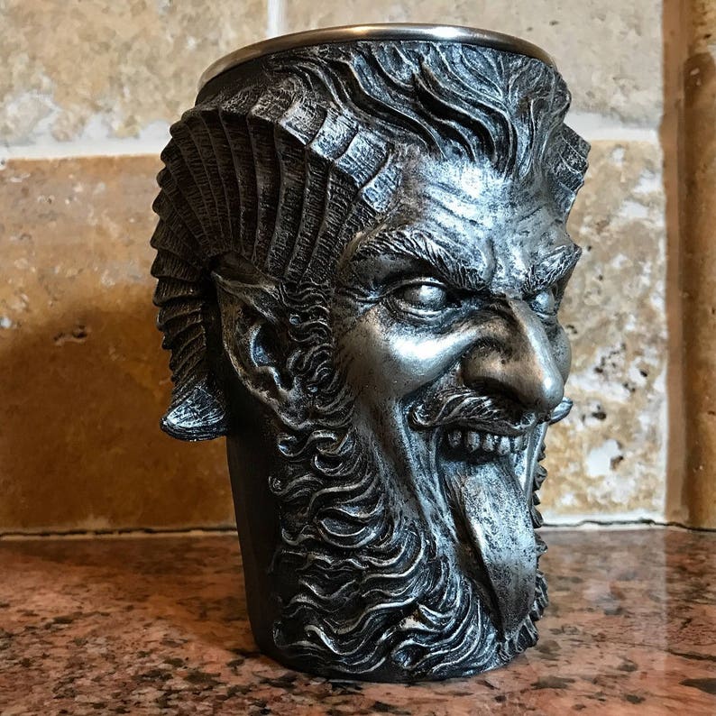 May include: A silver metal drinking cup shaped like a demon's head with a long tongue sticking out. The cup has a detailed design with a beard and horns.