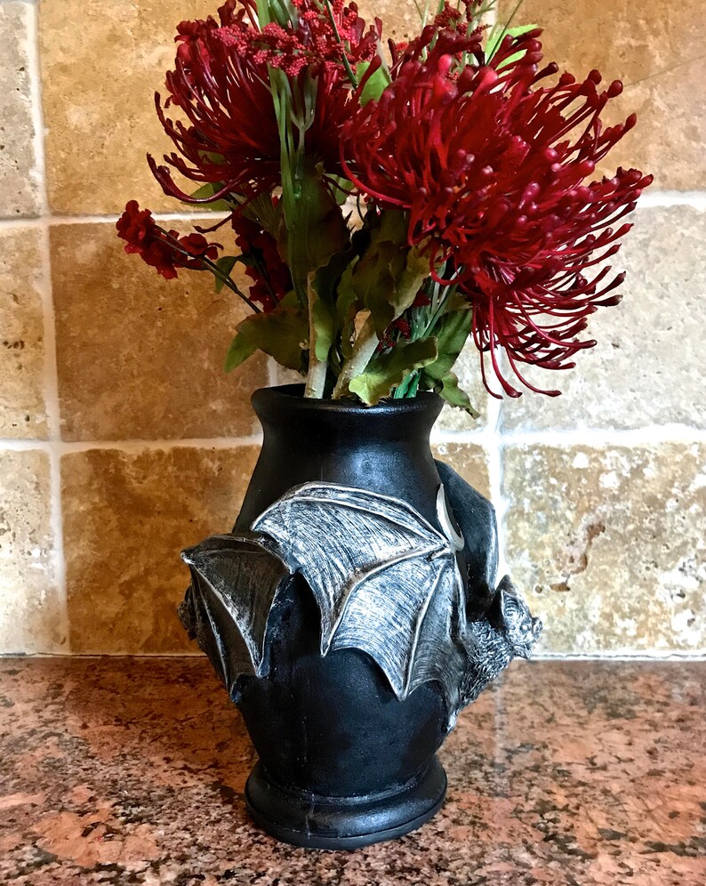 May include: Black ceramic vase with a bat design. The vase is filled with a bouquet of red flowers.