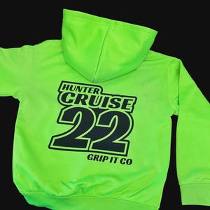 May include: A bright green hoodie with a hood and long sleeves. The back of the hoodie displays the text "HUNTER CRUISE 22 GRIP IT CO" in black block letters. The hoodie is made of a soft material.