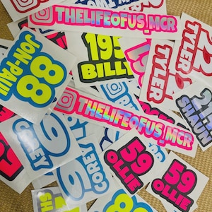 May include: A pile of stickers with diverse text and graphic designs. The stickers display names, numbers, and phrases in bold fonts, with bright colours such as pink, blue, yellow, and white. Some stickers incorporate social media icons.