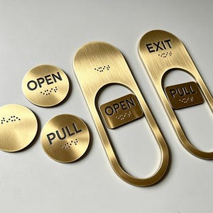 May include: A collection of brushed brass signage plates featuring black text and tactile braille. The set includes two circular discs labelled OPEN and PULL, and two elongated oval plates with cutouts, labelled OPEN and EXIT with PULL inserts.