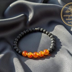 Può includere: Un braccialetto in pietra lavica nera con cinque perle ambra arancioni e rosse. Il braccialetto è esposto su un tessuto di raso grigio. L'immagine include un logo circolare dorato con la scritta "everything that adorns you".