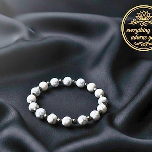 Puede incluir: Pulsera de cuentas blancas y grises con detalles plateados. La pulsera está hecha de cuentas redondas y veteadas y se muestra sobre una superficie de satén negro. Un logotipo circular dorado con el texto "everything that adorns you" está en la esquina superior derecha.
