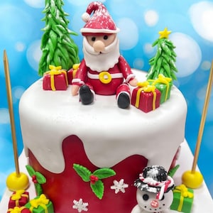 Puede incluir: Un pastel navideño festivo decorado con una figura de Papá Noel, árboles de Navidad verdes y cajas de regalo envueltas. El pastel tiene una parte superior blanca y una base roja, adornada con acebo y detalles de copos de nieve. Dos velas doradas están colocadas en el pastel.