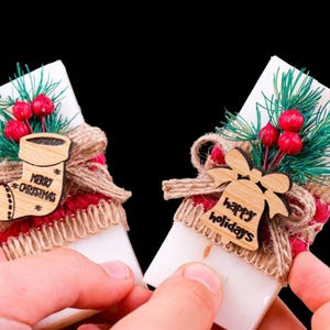 Puede incluir: Dos adornos navideños. Cada uno presenta una base rectangular blanca, adornada con bayas rojas, ramitas verdes y un recorte de madera. Uno dice "Merry Christmas" y el otro "happy holidays".