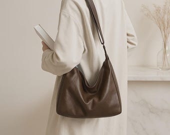 May include: A brown leather shoulder bag with a curved design. The bag has a long strap and is worn over the shoulder. The bag is a solid colour and appears to be made of a soft material. The bag is a neutral colour and can be used for everyday use.